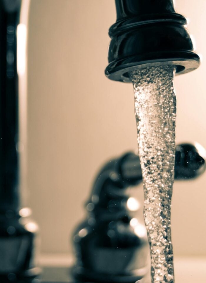 Close-up of a bathroom faucet running water, with a steady stream pouring from a dark metal tap while the handles and background remain softly blurred.