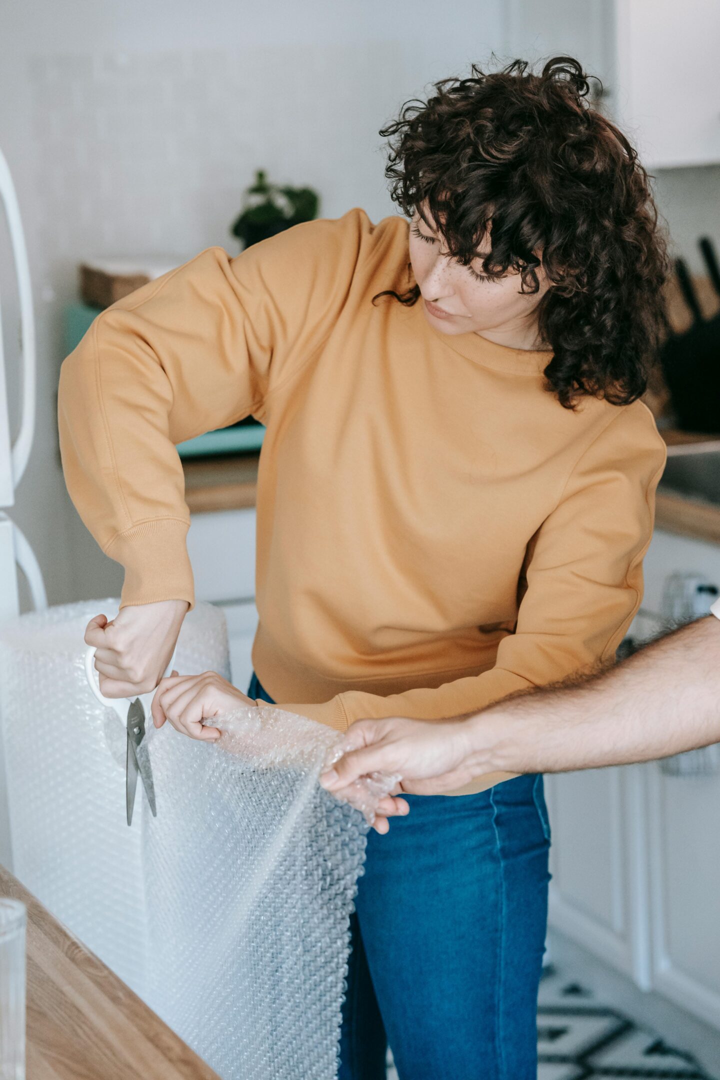 Woman cutting bubble wrap while packing items in a modern kitchen with wooden countertop.