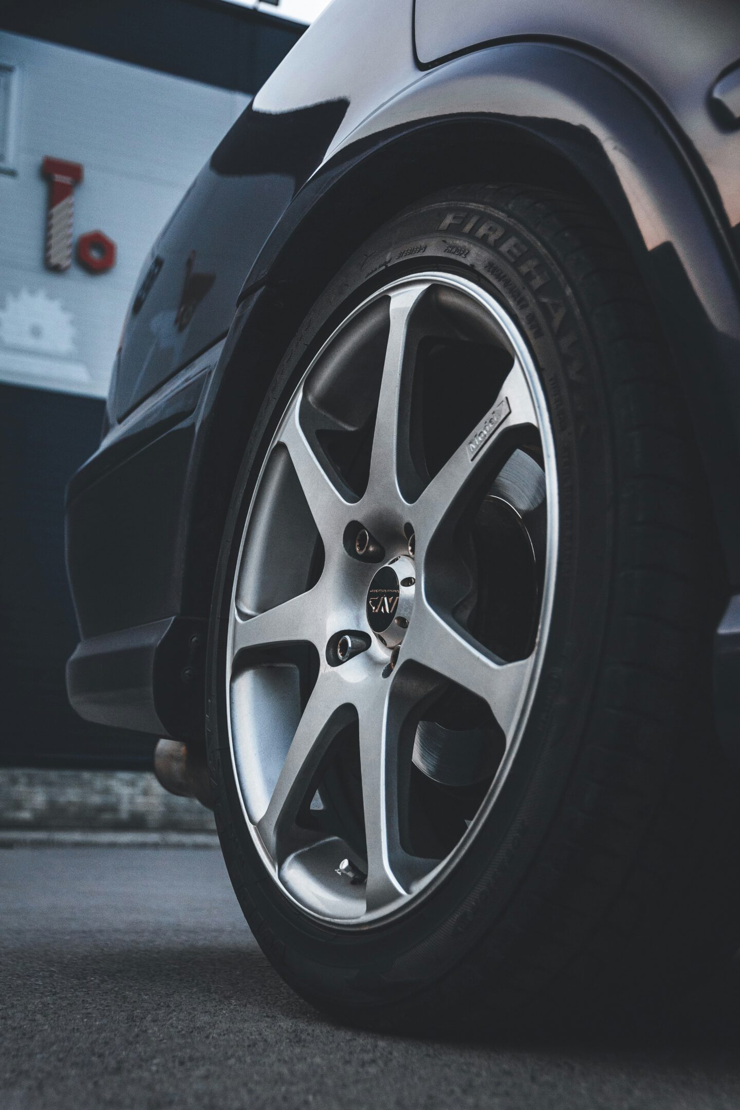 Close-up of a silver alloy car wheel with Firehawk tire mounted on a black vehicle, showing detailed rim spokes and tread pattern parked on pavement outside a garage.