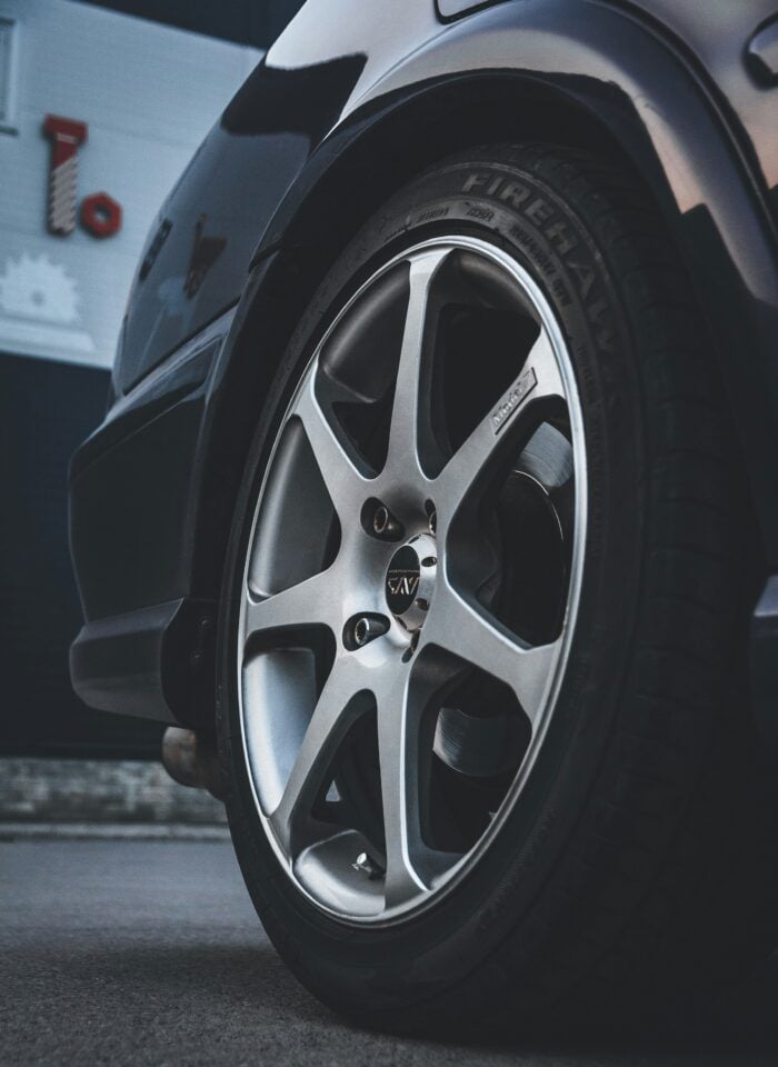 Close-up of a silver alloy car wheel with Firehawk tire mounted on a black vehicle, showing detailed rim spokes and tread pattern parked on pavement outside a garage.
