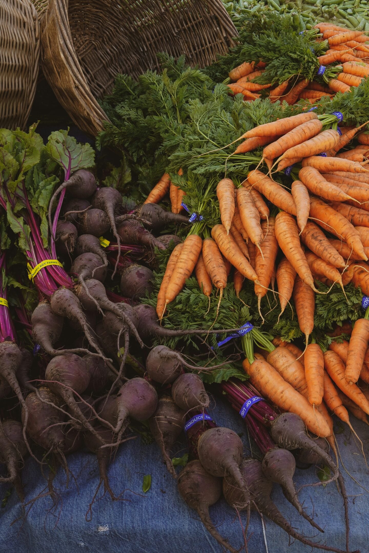Fresh carrots with leafy tops and dark purple beets piled together at a farmers market stall, displayed in rustic baskets on a wooden table.