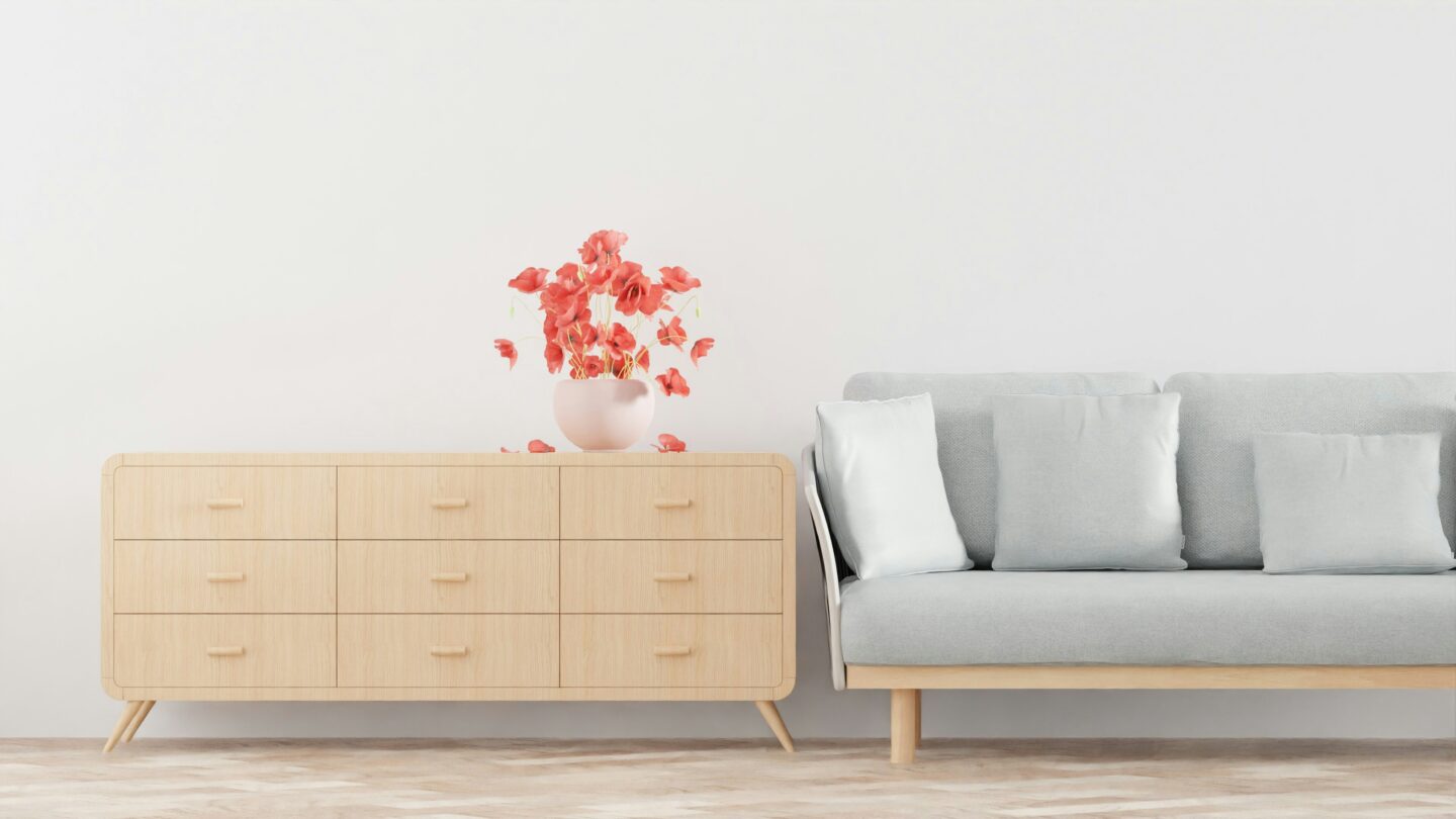 Minimalist living room with light wood sideboard, gray sofa, and vase of coral flowers on clean white wall background