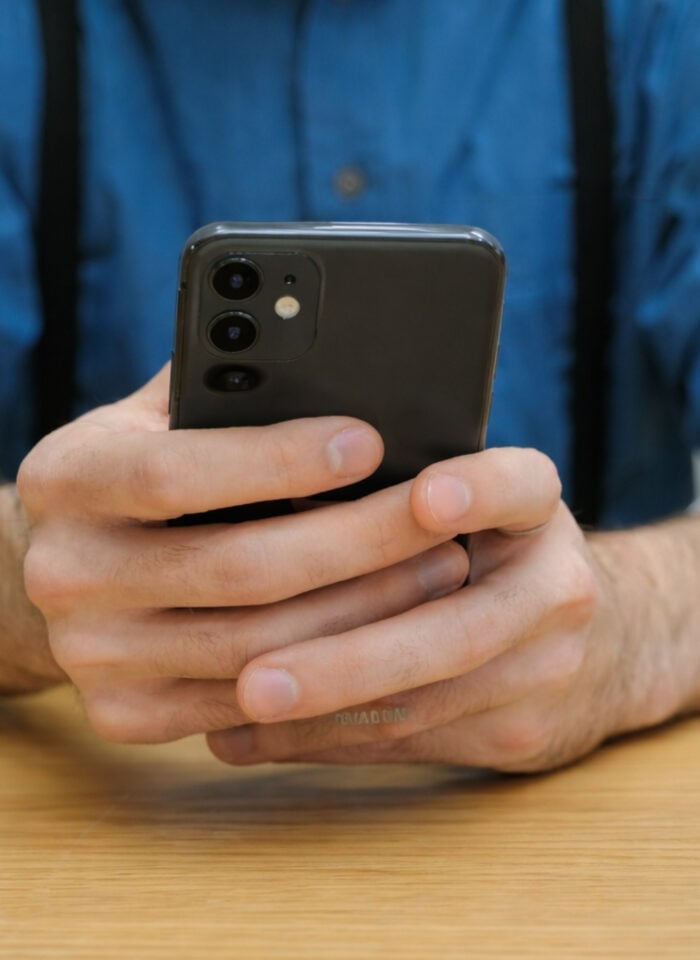 Person holding a modern smartphone with triple camera setup while sitting at a wooden table.