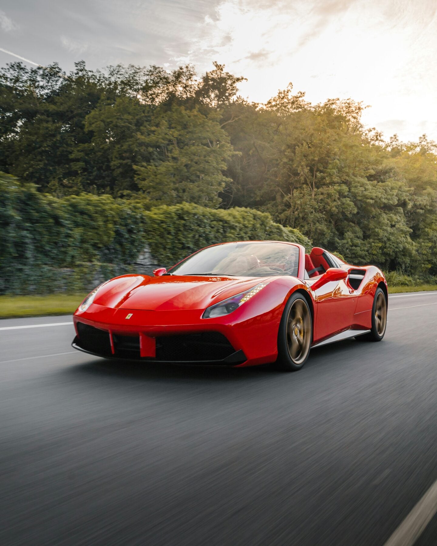 Red Ferrari 488 Spider convertible sports car driving on a scenic road surrounded by greenery, with motion blur and soft evening light highlighting its sleek, modern design