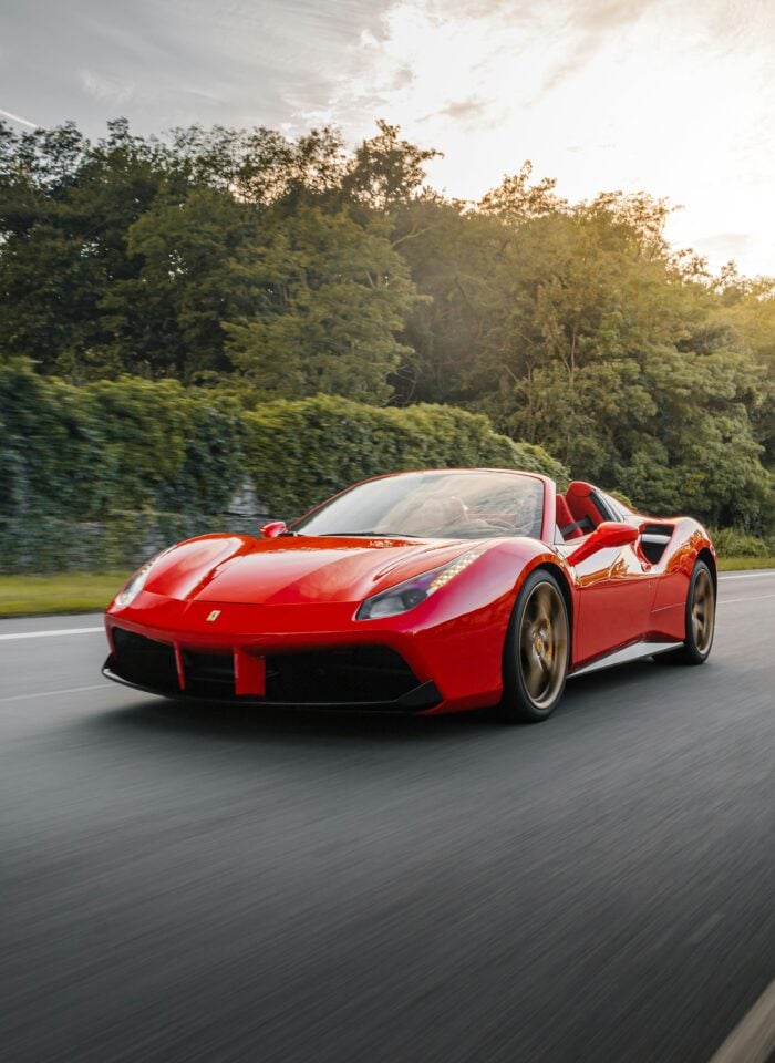 Red Ferrari 488 Spider convertible sports car driving on a scenic road surrounded by greenery, with motion blur and soft evening light highlighting its sleek, modern design