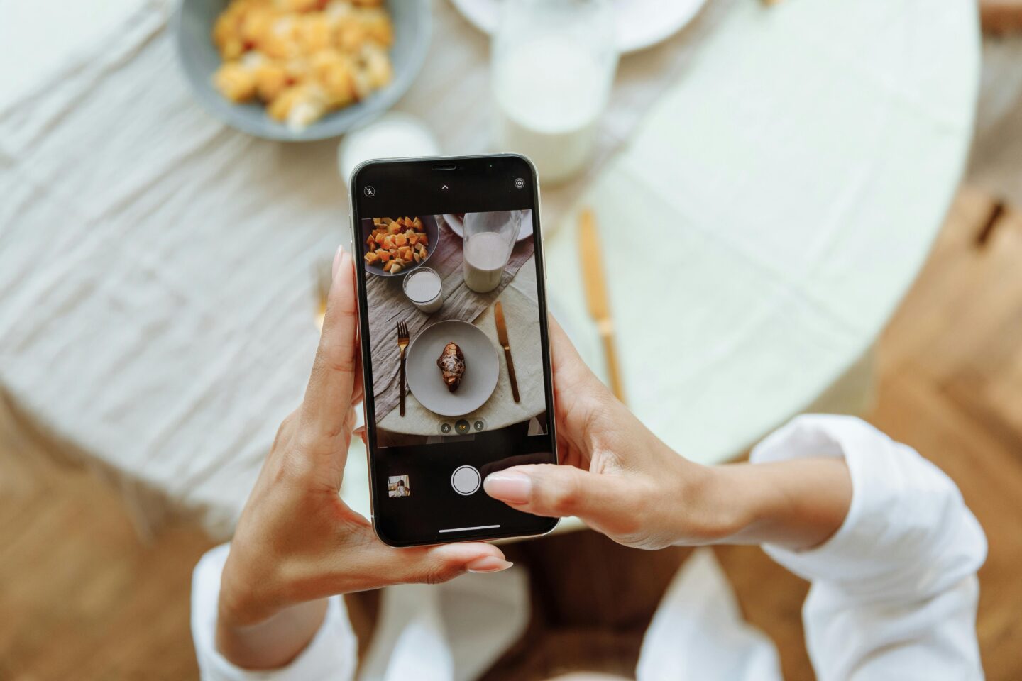 Person holding a smartphone and taking a photo of a plated breakfast on a marble table, with a croissant, milk, and fruit visible around the place setting.