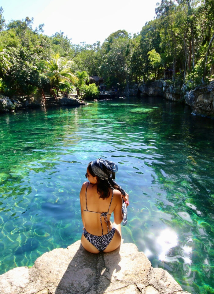 Woman sitting on rock at crystal clear cenote in Tulum Mexico surrounded by lush jungle swimming in natural freshwater sinkhole