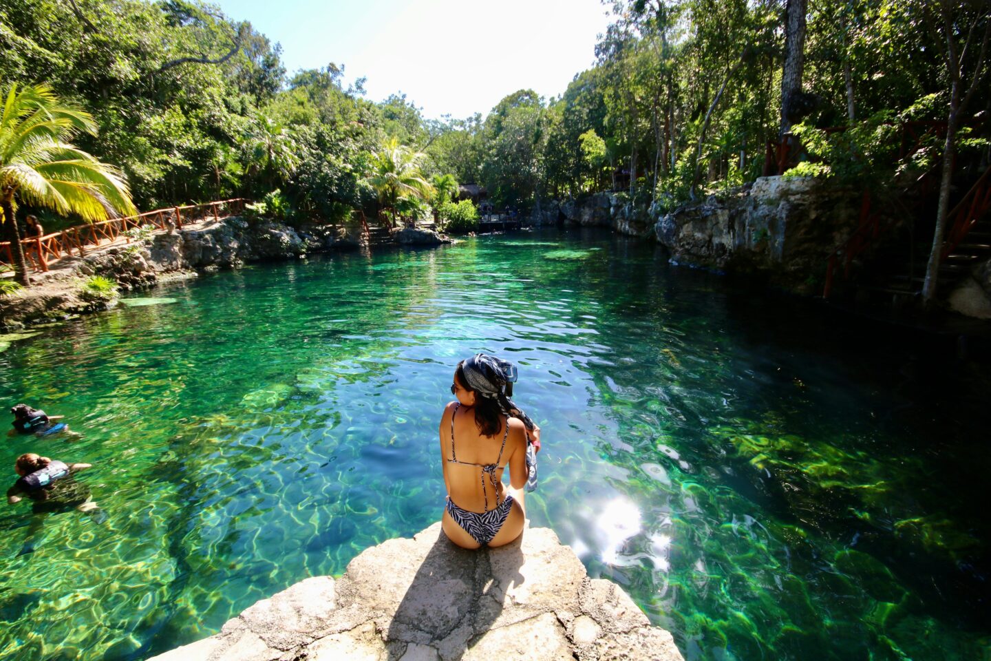 Woman sitting on rock at crystal clear cenote in Tulum Mexico surrounded by lush jungle swimming in natural freshwater sinkhole