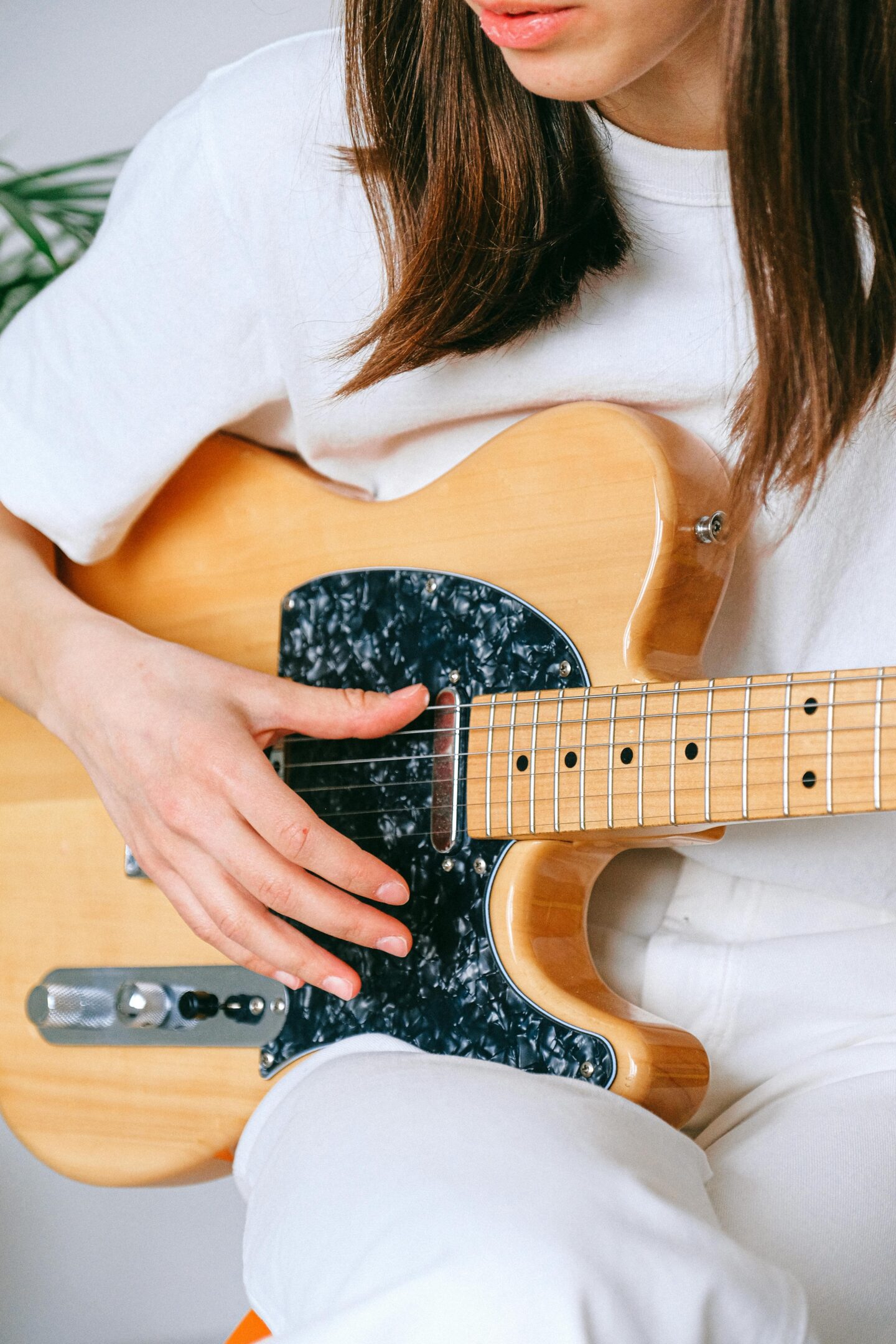 Young woman wearing a white outfit playing a natural wood electric guitar, close-up of hands strumming the strings and guitar body with a black pickguard.