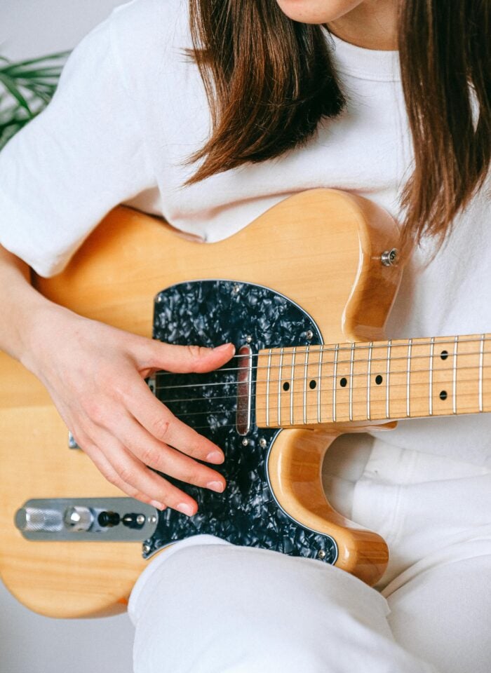 Young woman wearing a white outfit playing a natural wood electric guitar, close-up of hands strumming the strings and guitar body with a black pickguard.