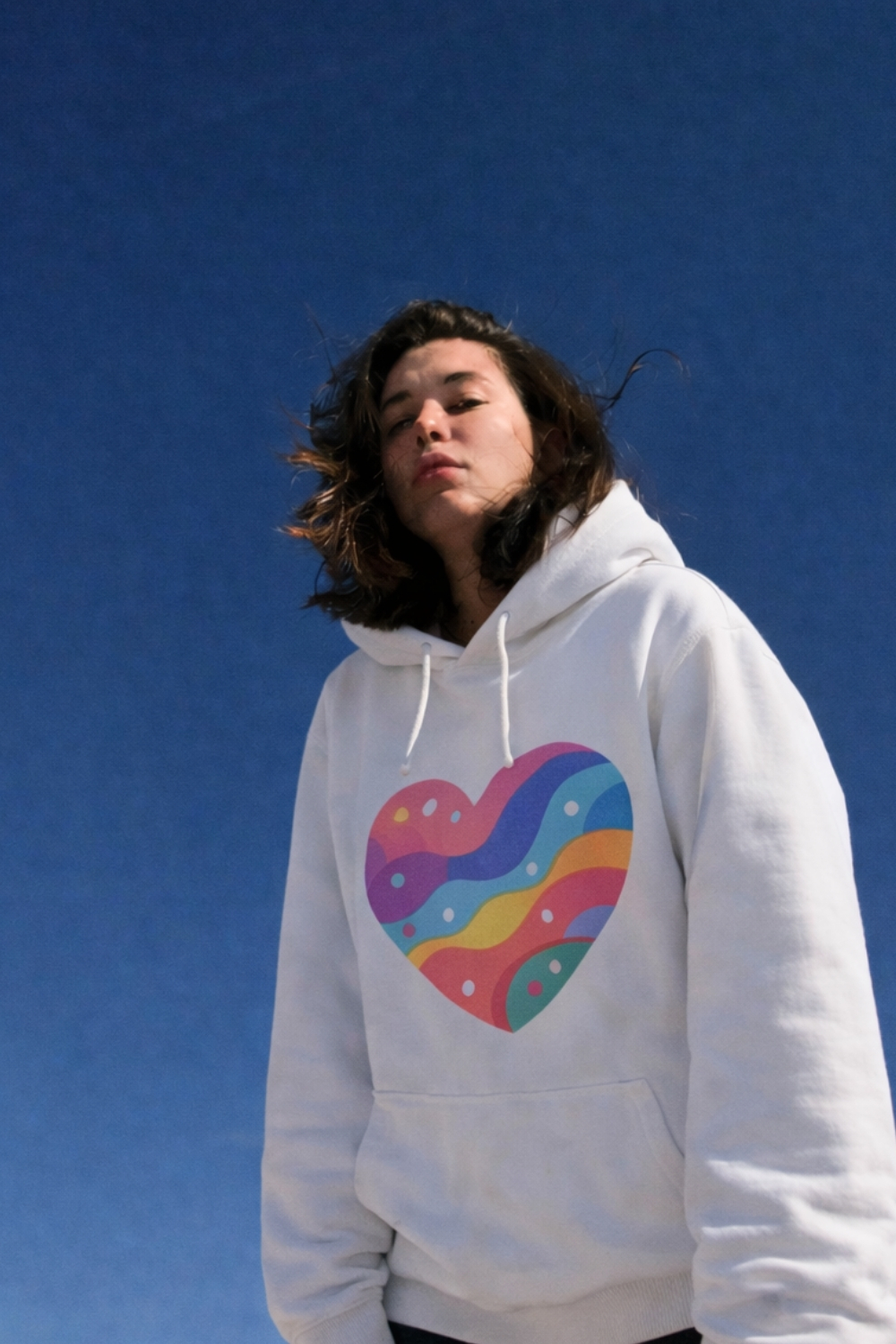 Young woman wearing a white hoodie with a colorful abstract heart design on the front, standing outdoors under a clear blue sky with wind-blown hair and a minimalist background.