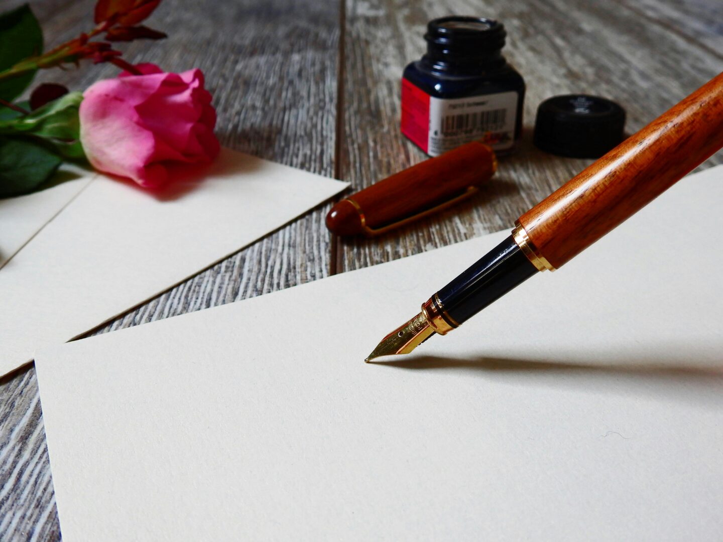Wooden fountain pen with a gold nib resting on blank cream paper, beside a pink rose, envelope, and open ink bottle on a rustic wooden table.