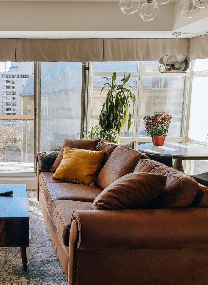 Bright, sunlit living room with large floor-to-ceiling windows, beige roman shades, and a warm-toned brown leather sofa styled with mustard and neutral cushions, paired with a dark wood coffee table on a textured rug, indoor plants, and a modern fireplace enhancing natural light in a contemporary apartment.