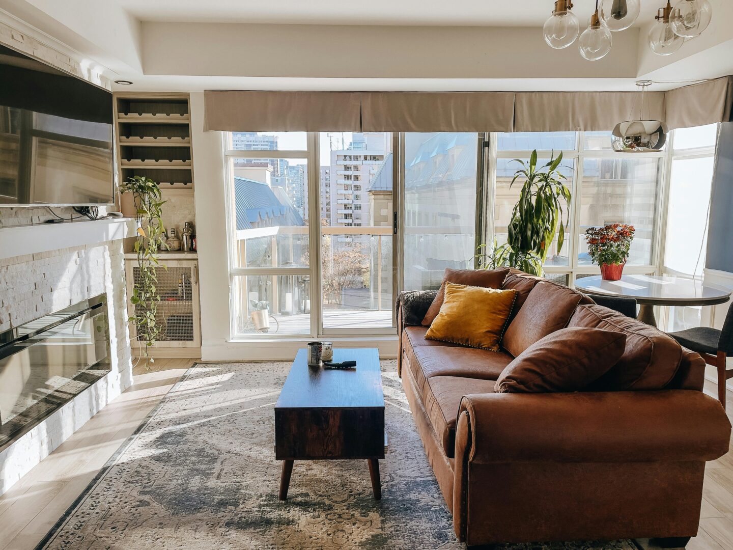Bright, sunlit living room with large floor-to-ceiling windows, beige roman shades, and a warm-toned brown leather sofa styled with mustard and neutral cushions, paired with a dark wood coffee table on a textured rug, indoor plants, and a modern fireplace enhancing natural light in a contemporary apartment.