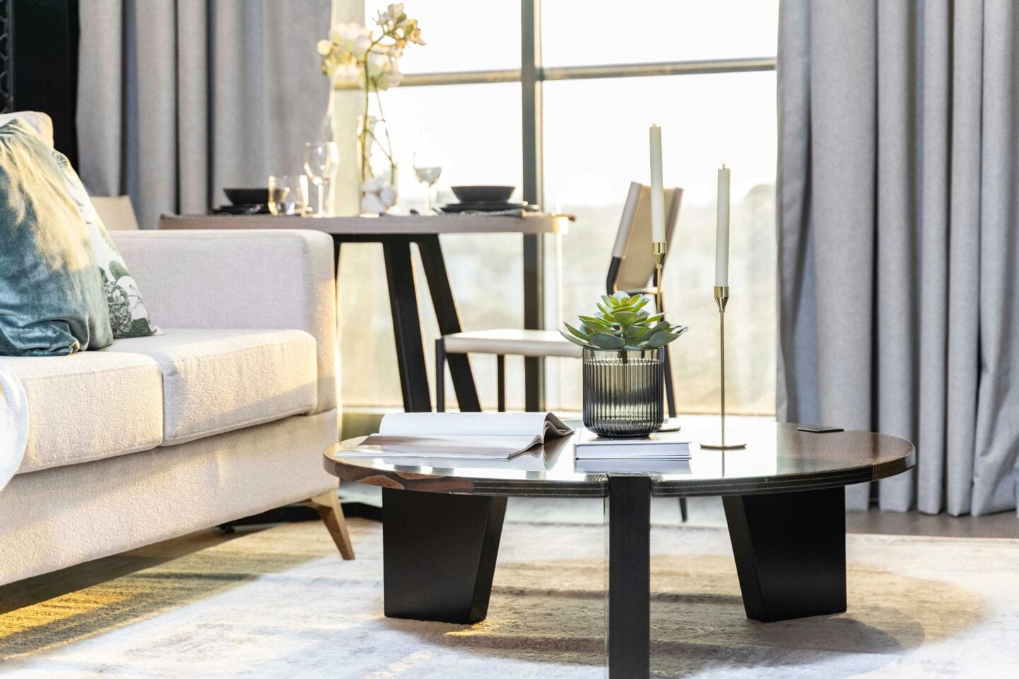Modern living room with soft natural sunlight streaming through tall windows framed by light grey curtains, featuring a neutral fabric sofa, round wooden coffee table with candles and a small plant, minimalist decor, and a cozy, airy apartment aesthetic.