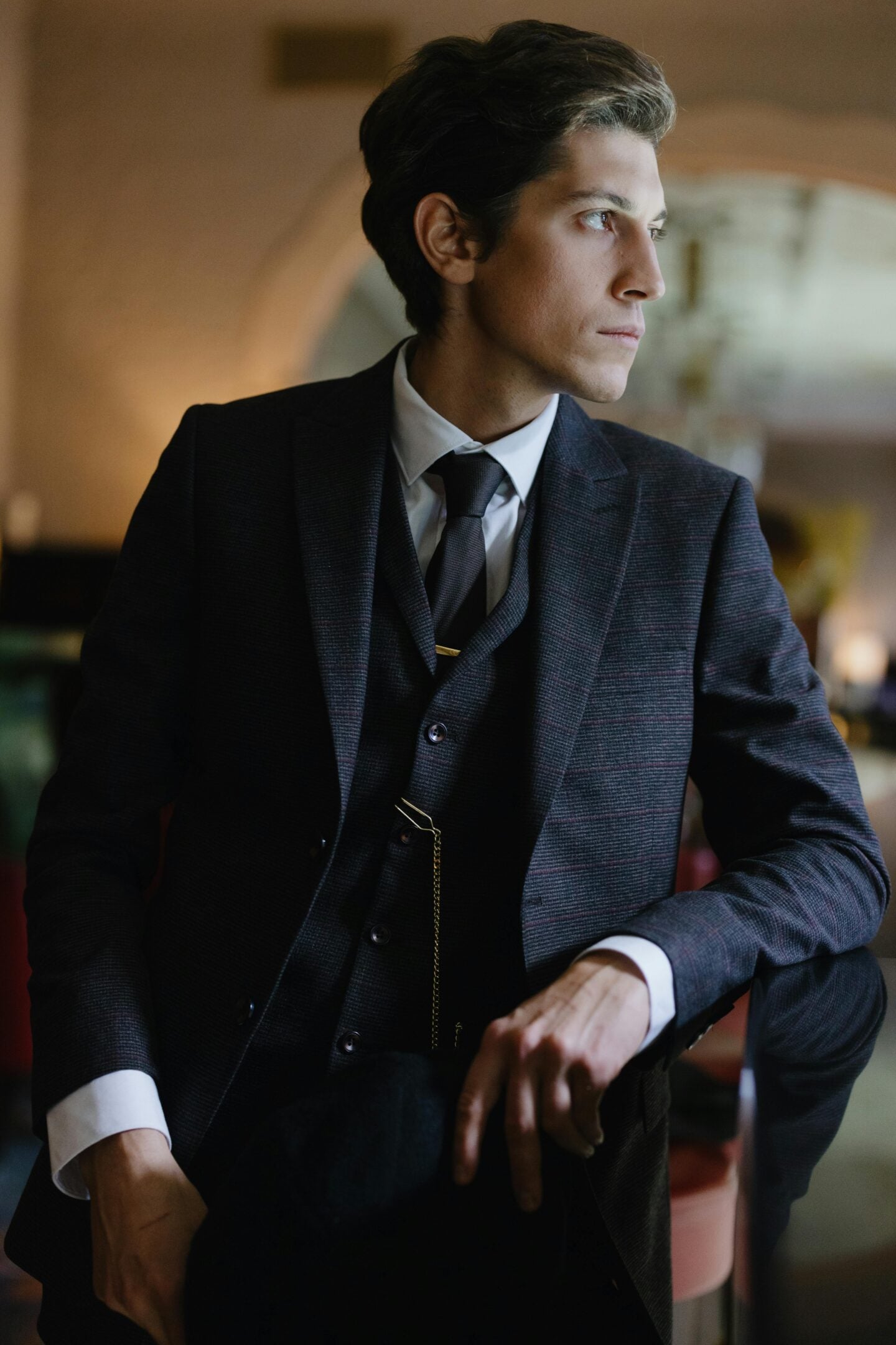 Elegant man in a tailored three-piece suit with tie and pocket watch chain, seated indoors under warm ambient lighting, looking to the side in a refined, classic setting.