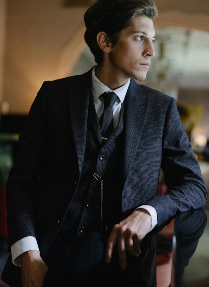 Elegant man in a tailored three-piece suit with tie and pocket watch chain, seated indoors under warm ambient lighting, looking to the side in a refined, classic setting.