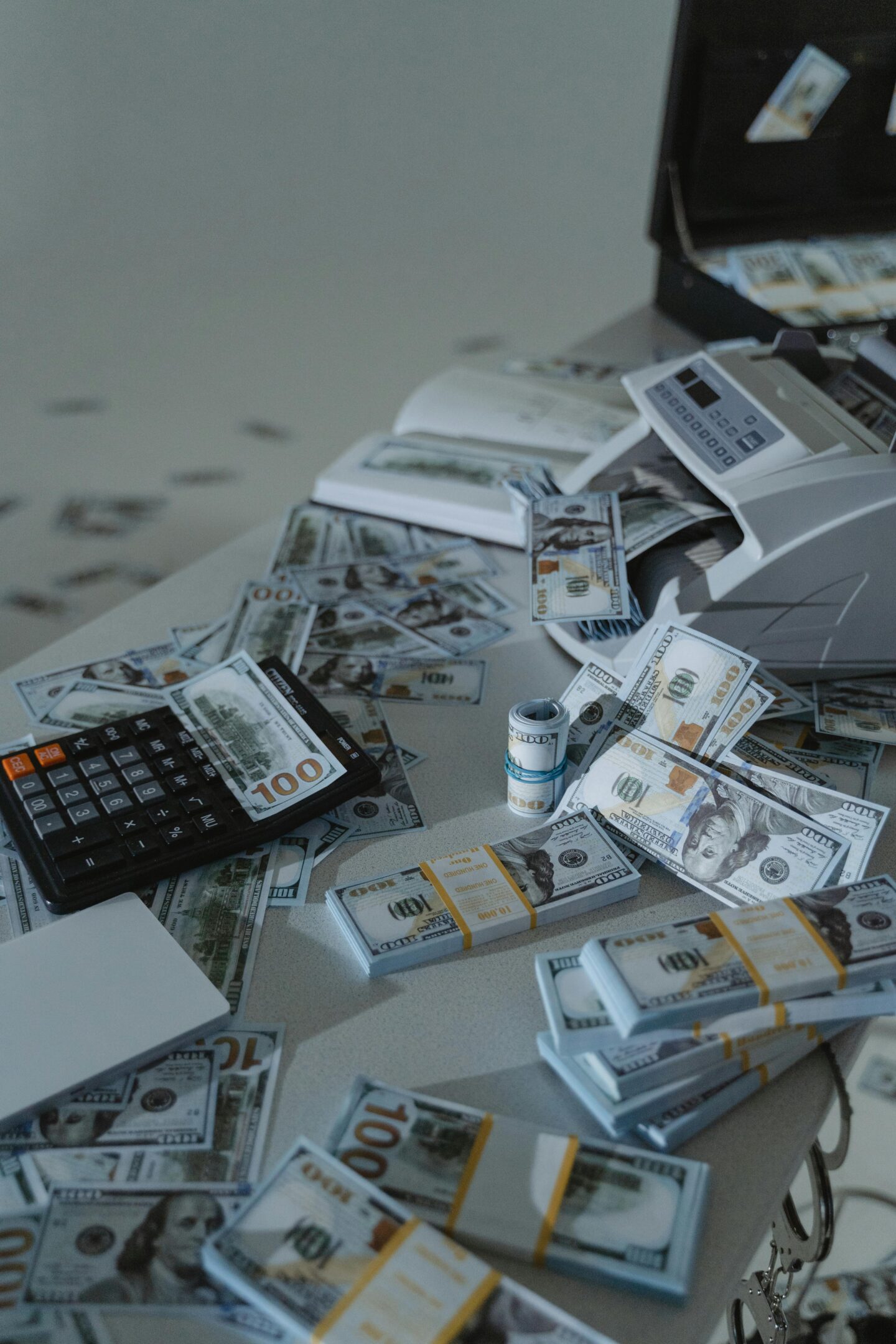 Stacks of US dollar bills scattered across a table with a money counting machine and calculator.