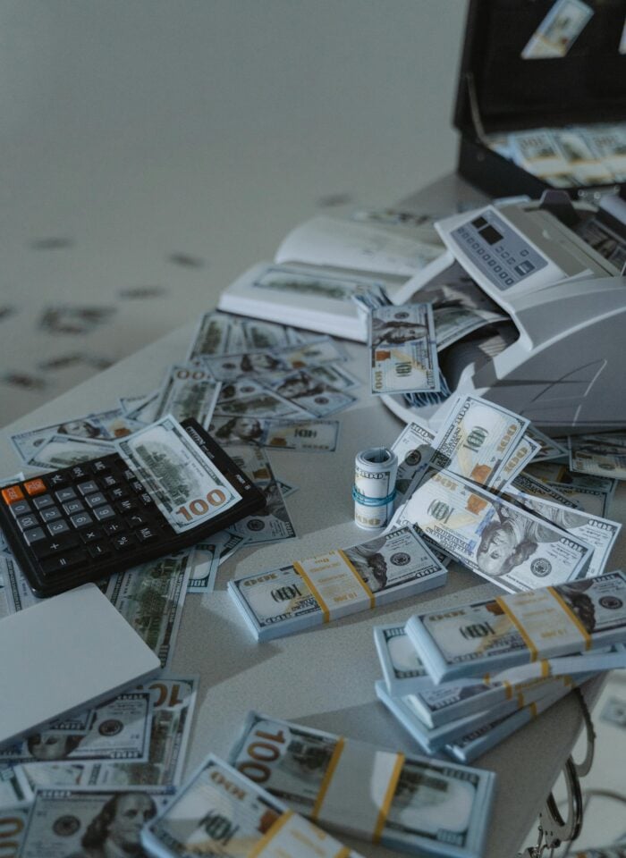Stacks of US dollar bills scattered across a table with a money counting machine and calculator.