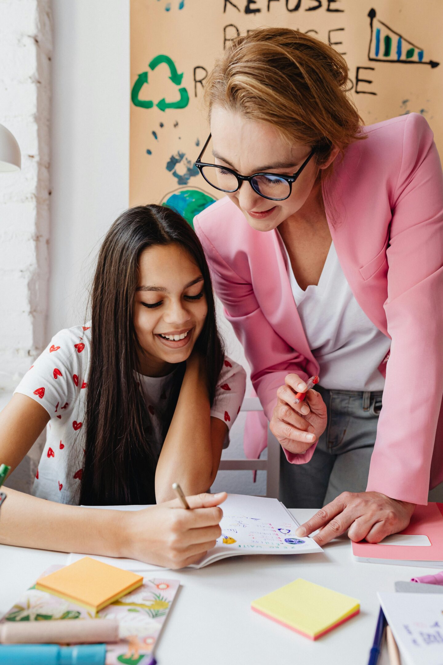 Teacher in a pink blazer helping a smiling student with schoolwork at a desk.