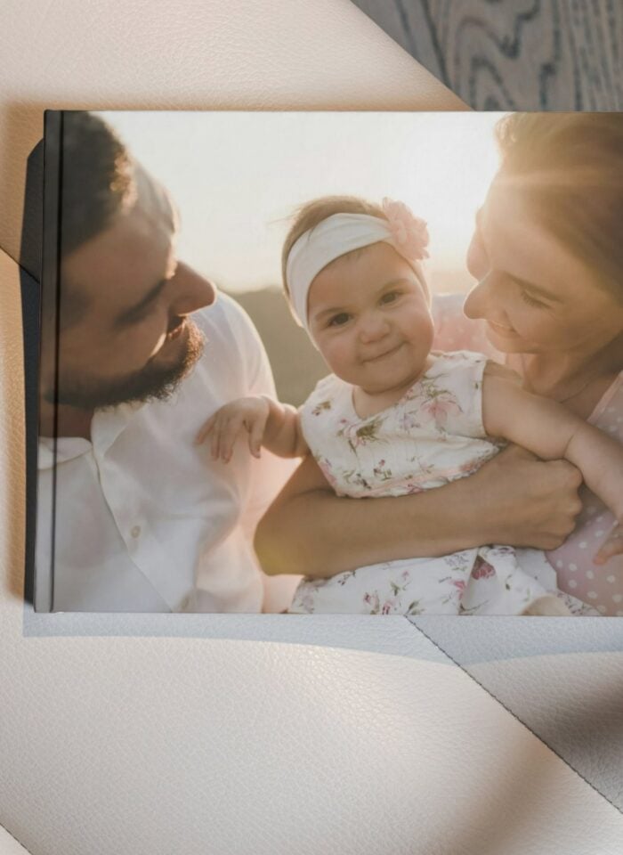 Photobook with a hardcover featuring a warm, sunlit family portrait of parents holding their smiling baby, resting on a soft leather surface.