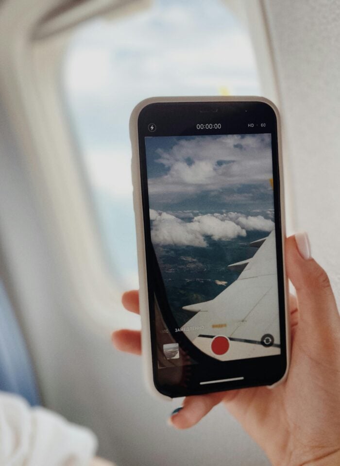 Person holding a smartphone recording a view of clouds and airplane wing through a plane window, capturing travel footage mid-flight.
