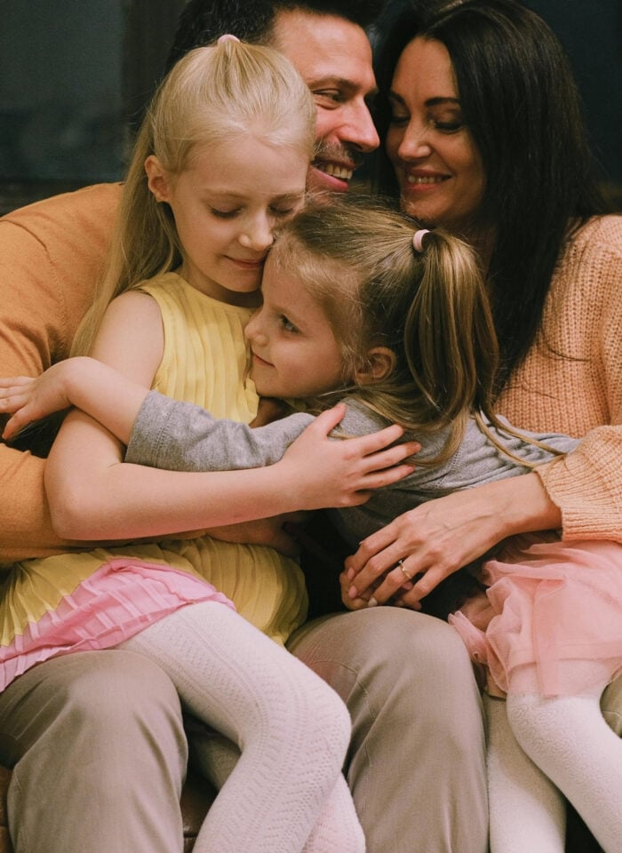 Warm family moment with parents and two young daughters cuddling together on a brown leather sofa, smiling and hugging in a cozy indoor setting.