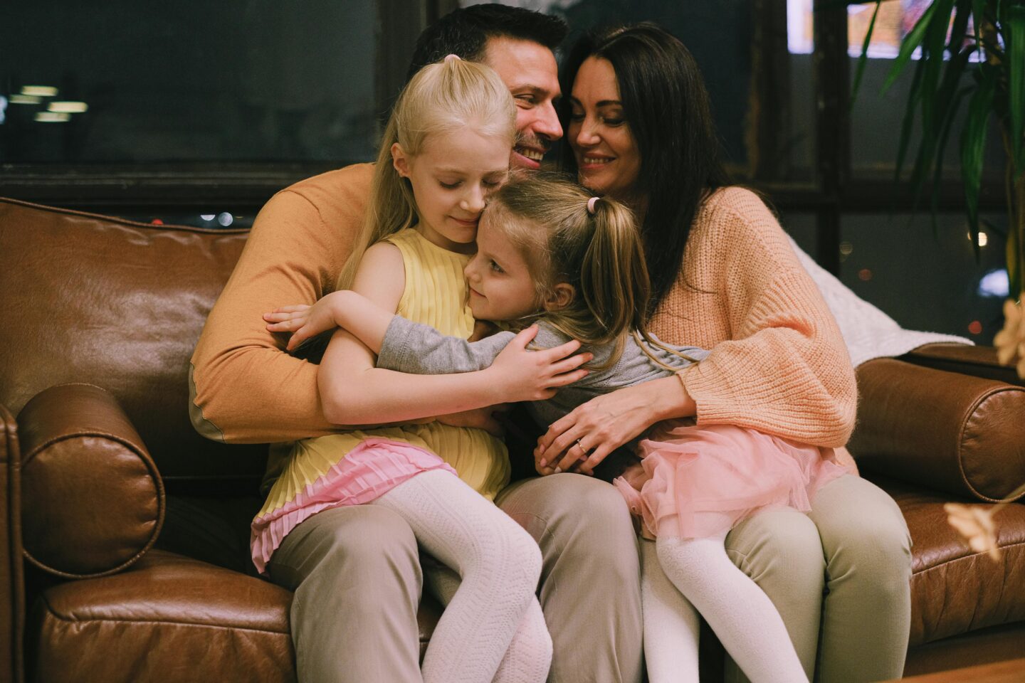 Warm family moment with parents and two young daughters cuddling together on a brown leather sofa, smiling and hugging in a cozy indoor setting.