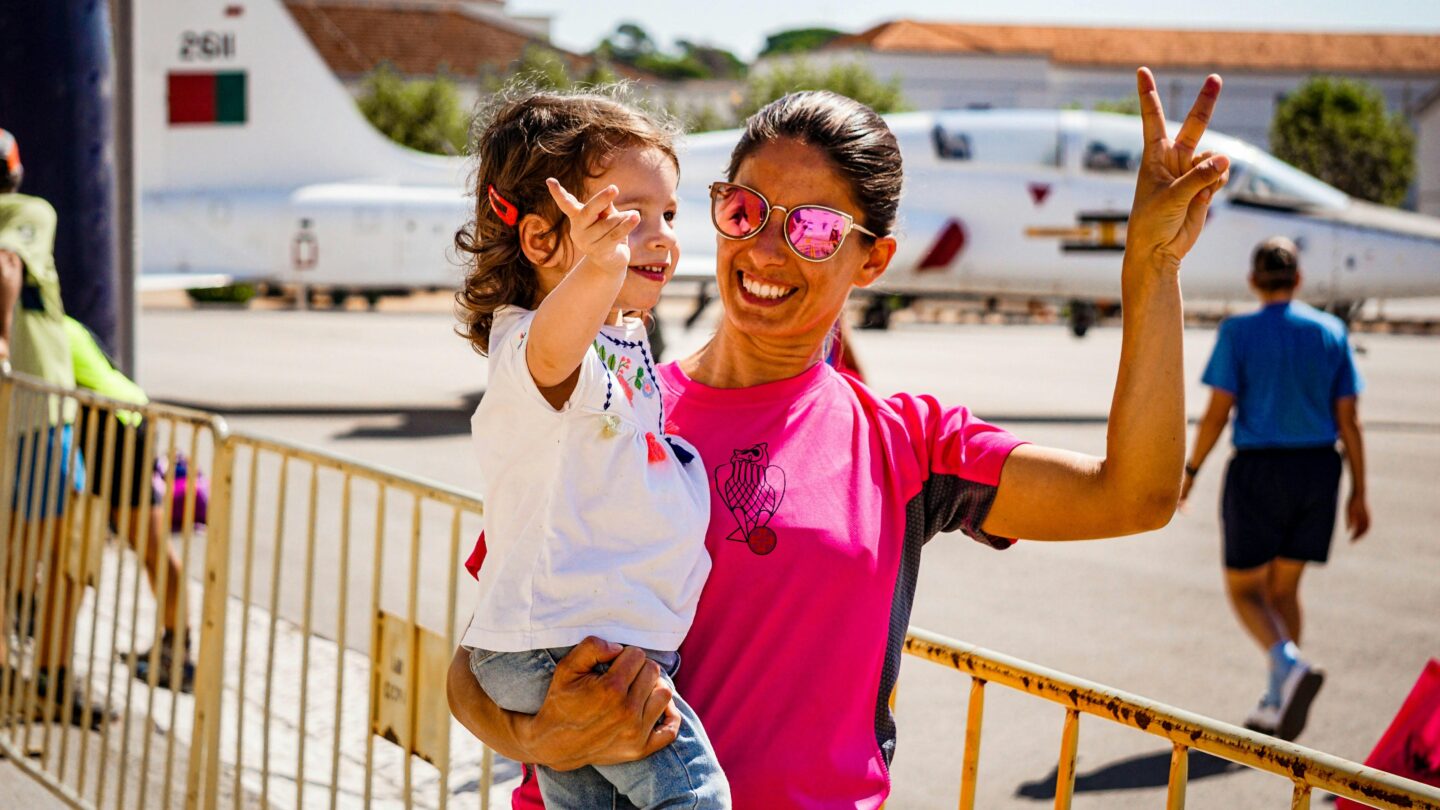 Smiling mother wearing pink shirt and reflective sunglasses holding a toddler at an airfield, both making peace signs with a small airplane in the background.