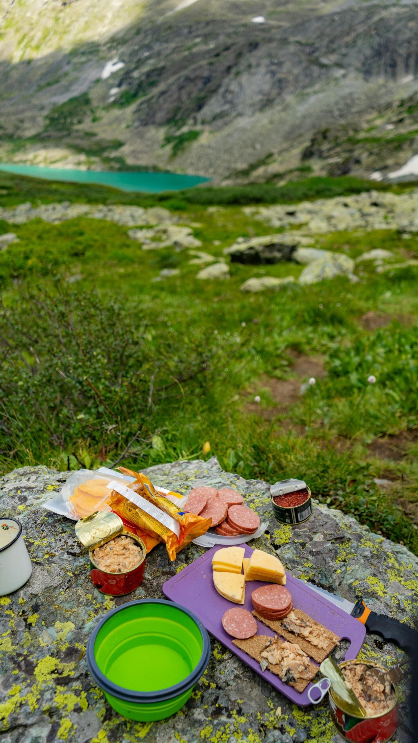 Hiking lunch spread with crackers, cheese, salami, and canned fish on a rock by a mountain lake, showcasing no-fridge hiking lunch ideas.