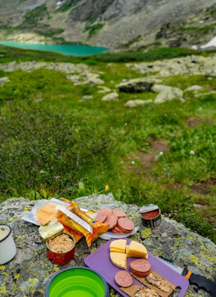 Hiking lunch spread with crackers, cheese, salami, and canned fish on a rock by a mountain lake, showcasing no-fridge hiking lunch ideas.