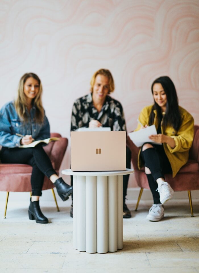 Three young adults sitting on pink armchairs in a modern office, writing in notebooks with a laptop on a small round table in front of them, against a soft abstract pastel wall background.