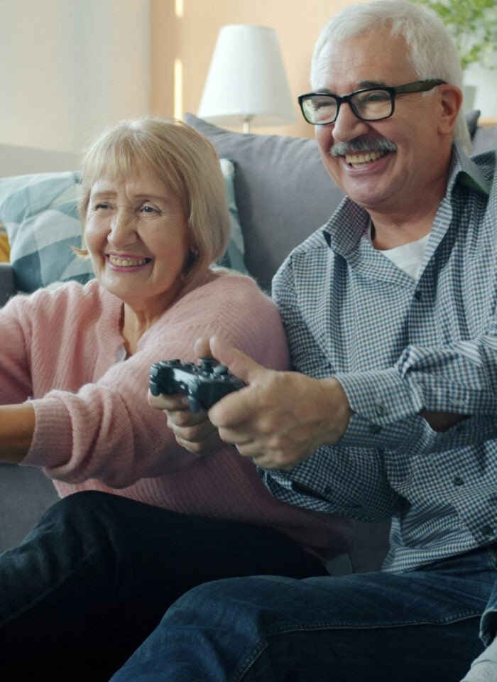 Smiling elderly couple sitting on the floor in a bright modern living room, holding game controllers and playing video games together, enjoying a fun and active moment at home with cozy decor and natural light.