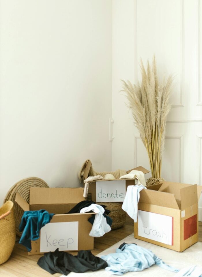 Minimalist corner of a bright room with cardboard boxes labeled keep, donate, and trash filled with clothes, showcasing home decluttering and organizing before a move, with neutral decor, woven baskets, and dried pampas grass in a soft, airy interior.