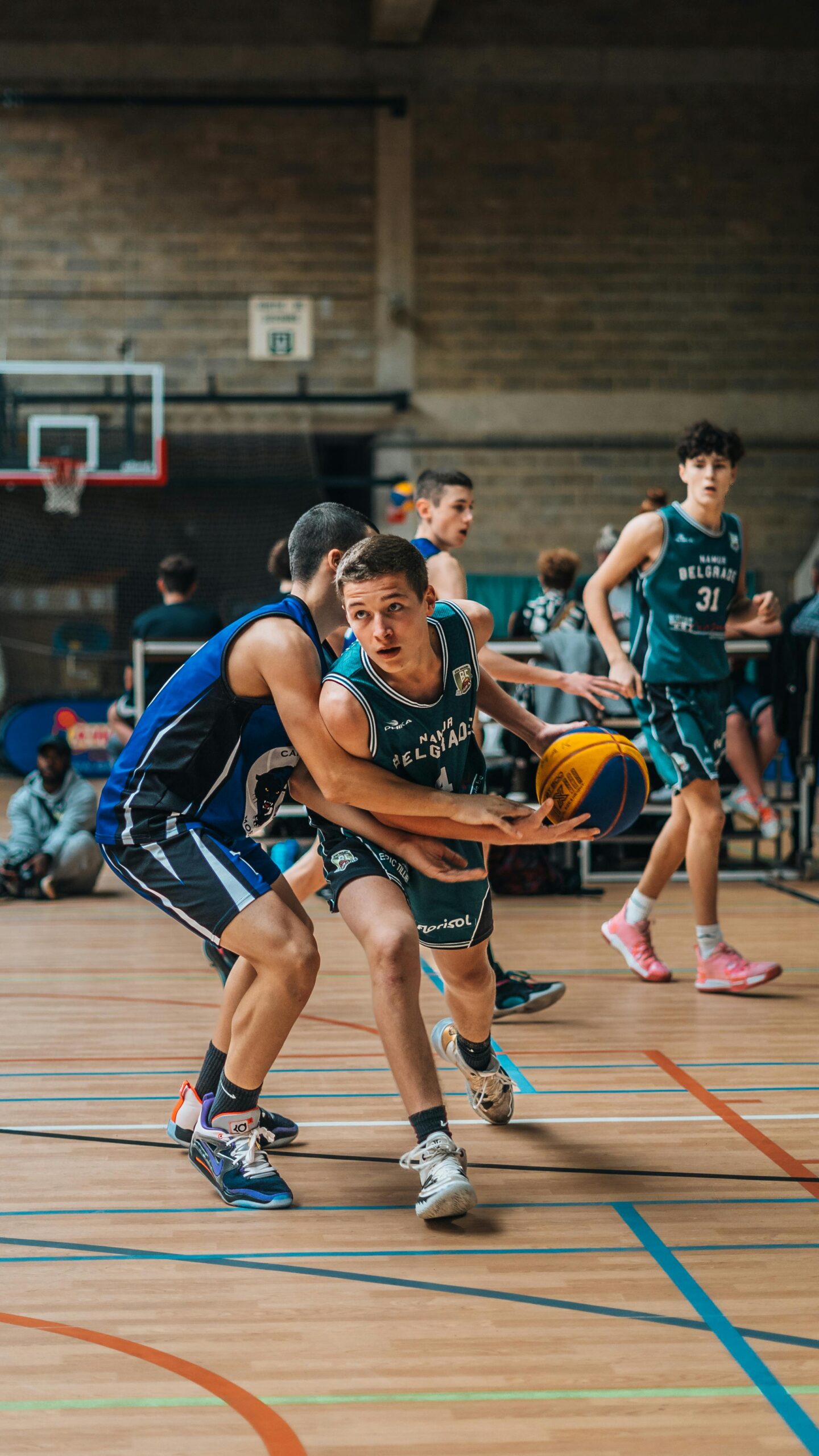 Youth basketball players competing in indoor game, athlete dribbling ball while defended, team sport action in gymnasium with dynamic movement and focus.