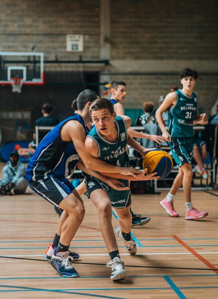 Youth basketball players competing in indoor game, athlete dribbling ball while defended, team sport action in gymnasium with dynamic movement and focus.