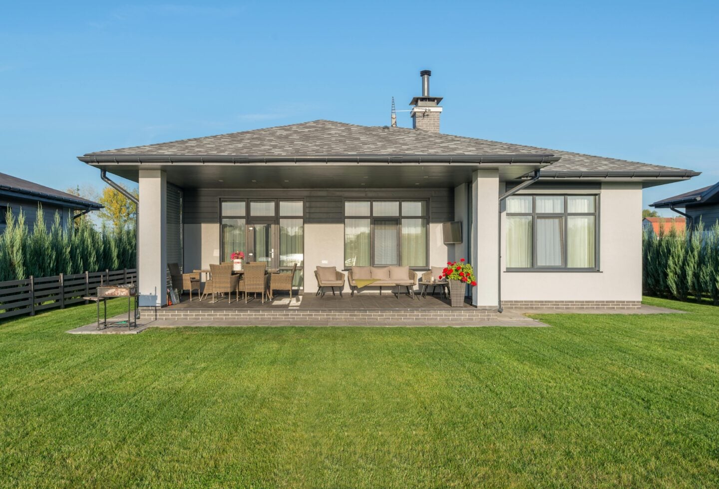 Modern single-story house with a covered patio featuring outdoor seating and dining furniture, set against a neatly maintained green lawn and clear blue sky.
