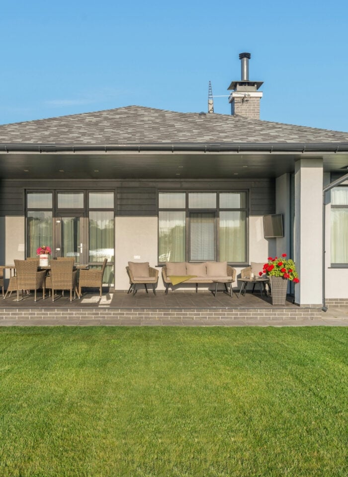 Modern single-story house with a covered patio featuring outdoor seating and dining furniture, set against a neatly maintained green lawn and clear blue sky.