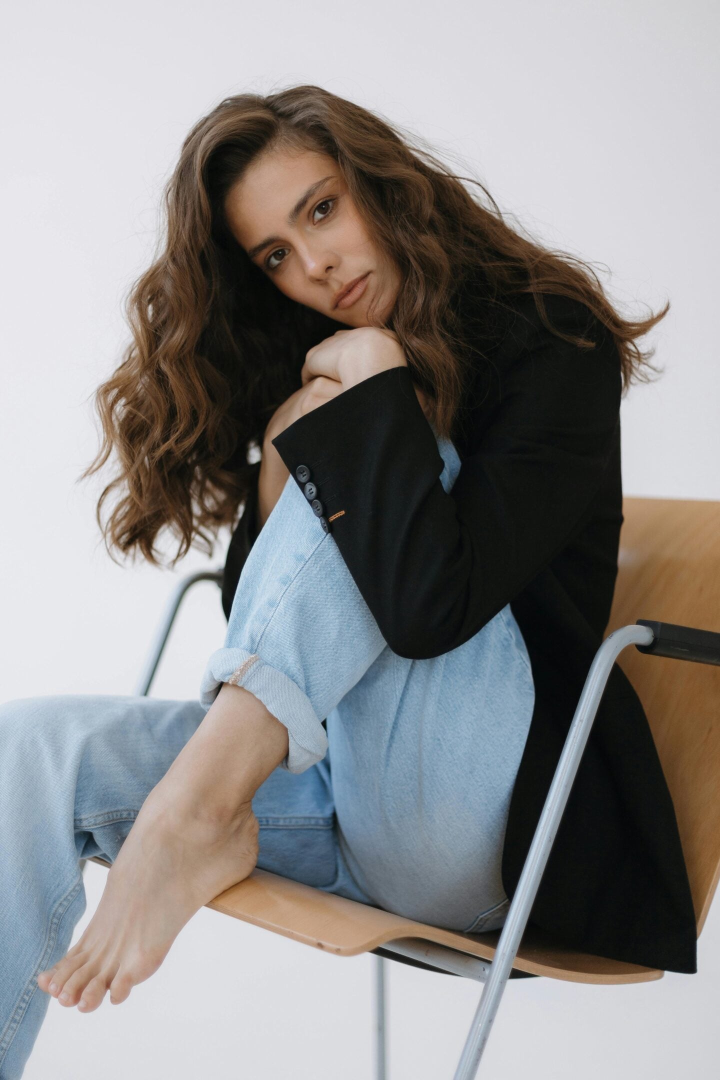 Woman with long wavy hair wearing a black blazer and light blue jeans sitting barefoot on a chair, posing in a relaxed yet confident way.