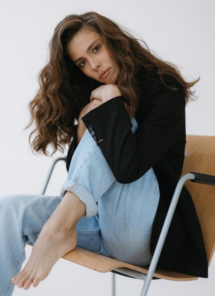 Woman with long wavy hair wearing a black blazer and light blue jeans sitting barefoot on a chair, posing in a relaxed yet confident way.