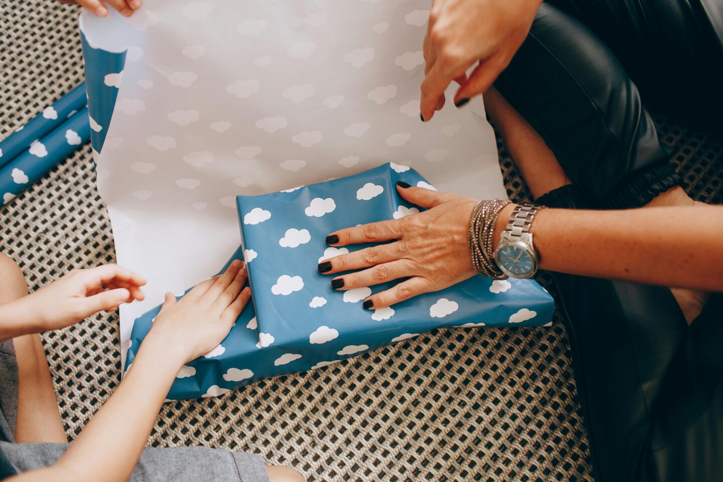 Person wrapping a gift in blue paper with white cloud pattern, hands preparing a cozy care package with child helping, thoughtful DIY gift wrapping at home.