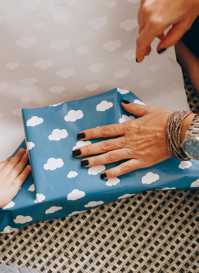Person wrapping a gift in blue paper with white cloud pattern, hands preparing a cozy care package with child helping, thoughtful DIY gift wrapping at home.