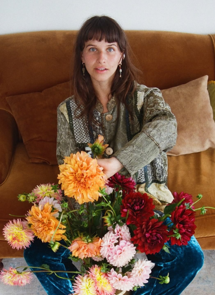 Woman sitting on a brown couch holding a colorful bouquet of dahlias in shades of orange, red, pink, and cream.