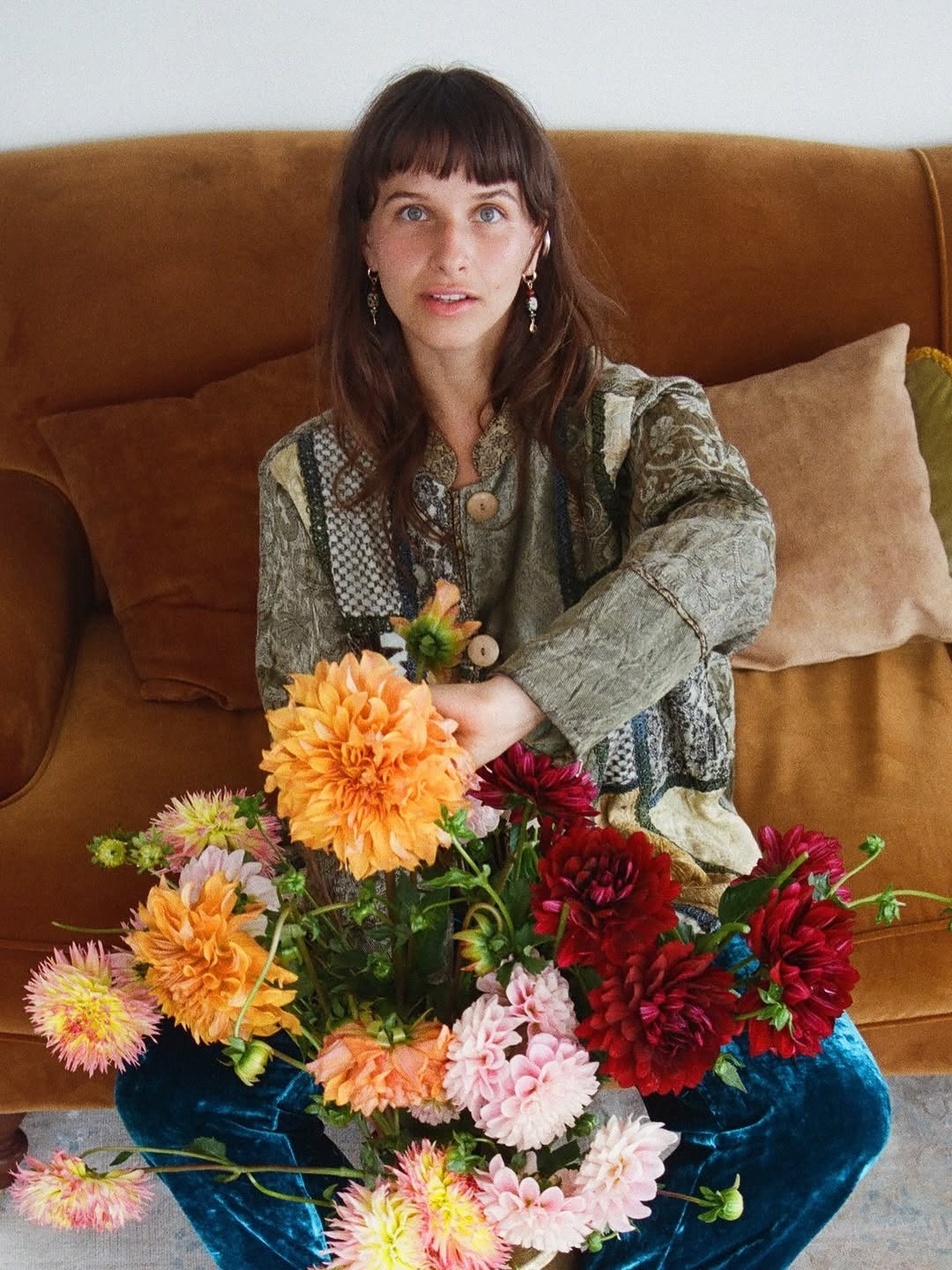 Woman sitting on a brown couch holding a colorful bouquet of dahlias in shades of orange, red, pink, and cream.