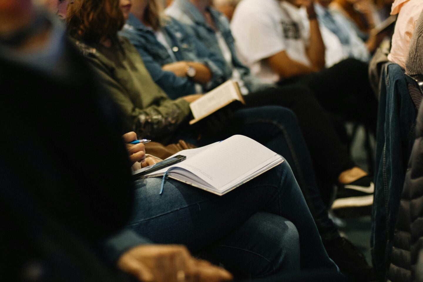 Students seated in a lecture or seminar, with one person holding a pen and writing notes in an open notebook on their lap while others sit nearby, some reading books, creating a focused academic atmosphere.