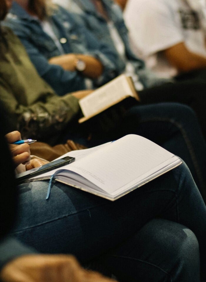 Students seated in a lecture or seminar, with one person holding a pen and writing notes in an open notebook on their lap while others sit nearby, some reading books, creating a focused academic atmosphere.