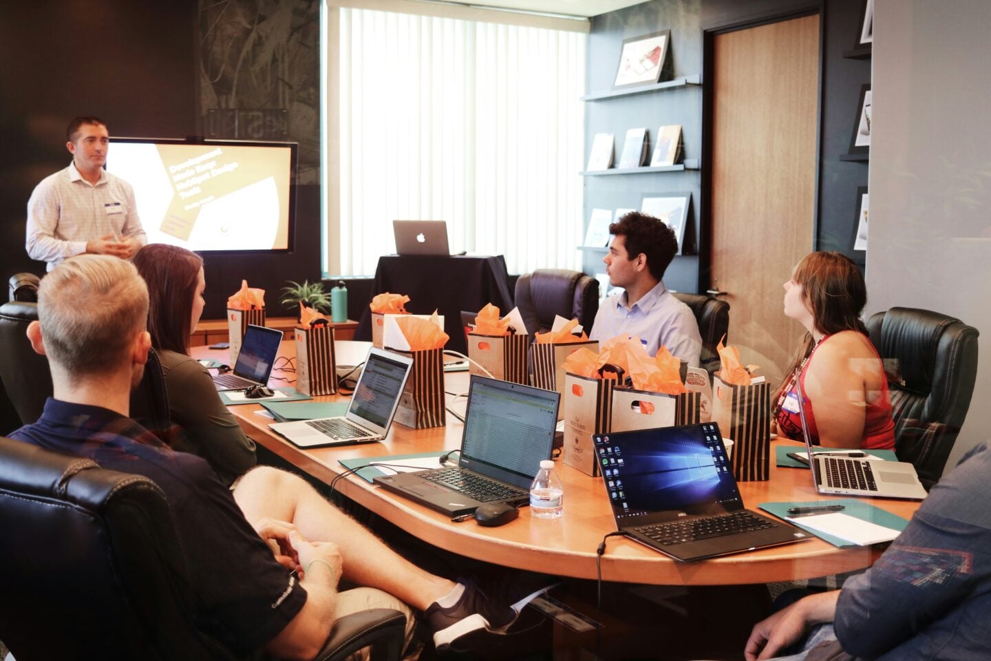 Business meeting in a modern conference room with a presenter giving a presentation on a screen while team members sit around a table with laptops, notepads, and gift bags, listening attentively.