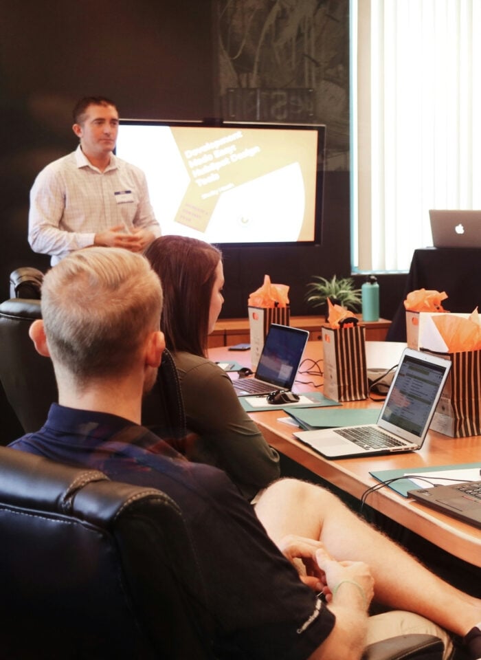 Business meeting in a modern conference room with a presenter giving a presentation on a screen while team members sit around a table with laptops, notepads, and gift bags, listening attentively.
