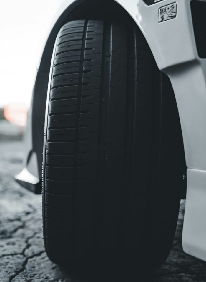 Close-up of a car tire with visible tread on a white vehicle, parked on a textured road surface, highlighting tire condition and performance for driving safety.