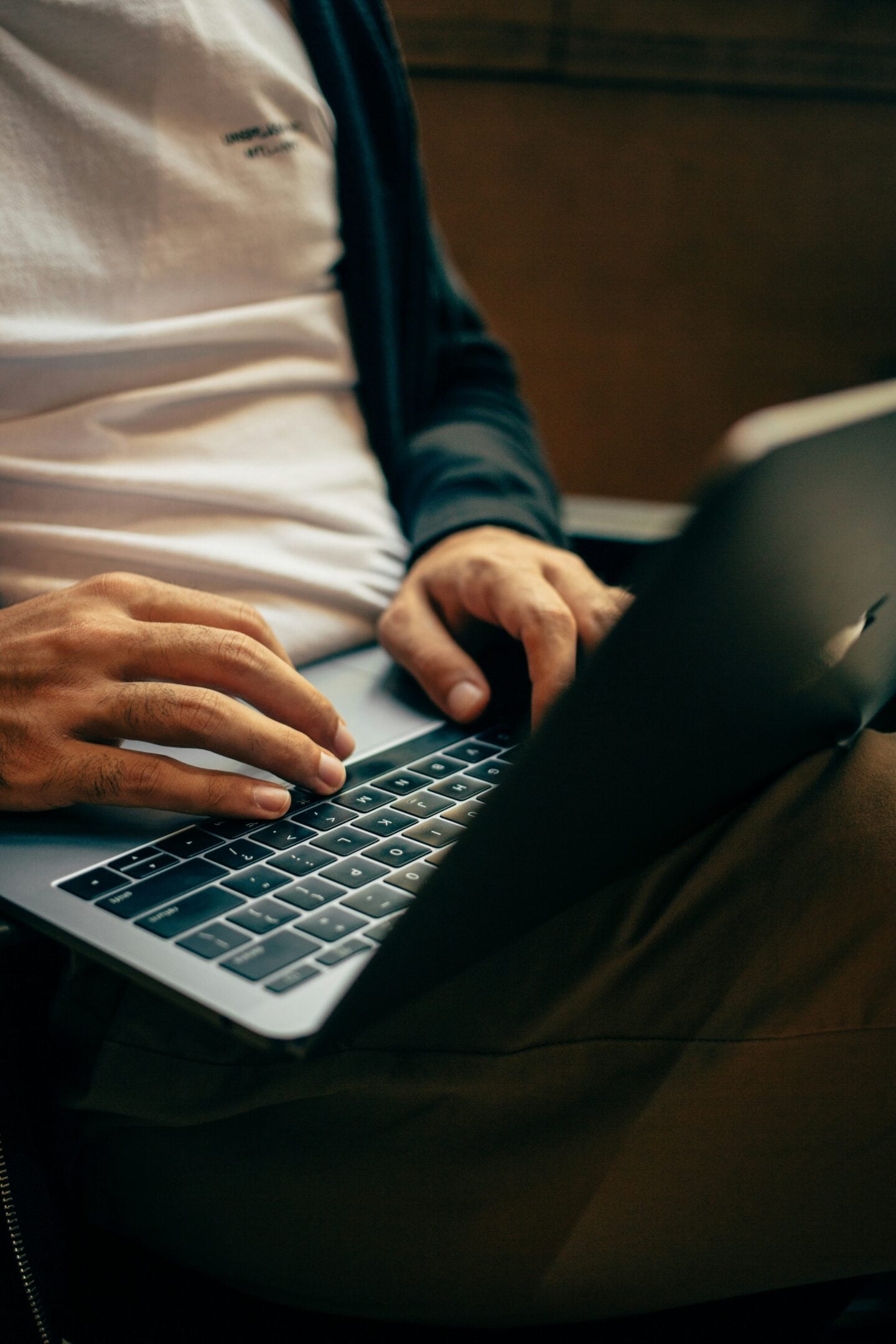 Close-up of a person typing on a laptop keyboard while sitting indoors, wearing a white t-shirt and dark cardigan, with a relaxed posture suggesting online browsing or shopping.